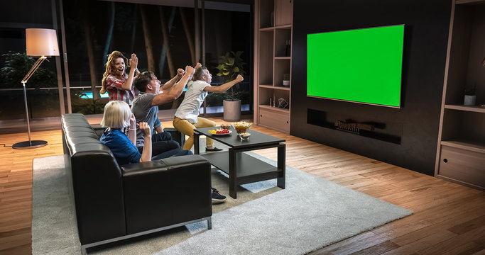A group of students is watching a TV and celebrating some joyful sports moment, sitting on the couch in the living room.