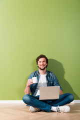 smiling man holding coffee cup and laptop and sitting on floor and looking at camera