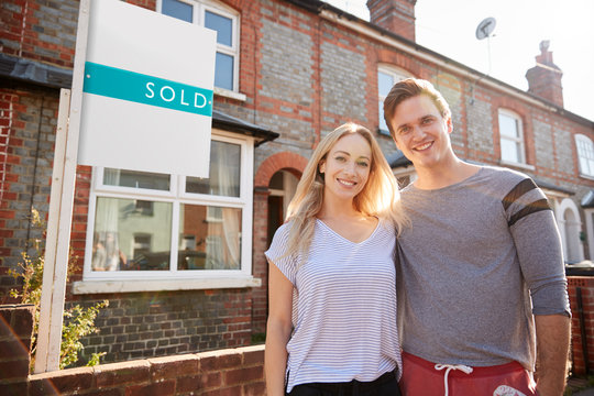 Portrait Of Excited Couple Standing Outside New Home With Sold Sign
