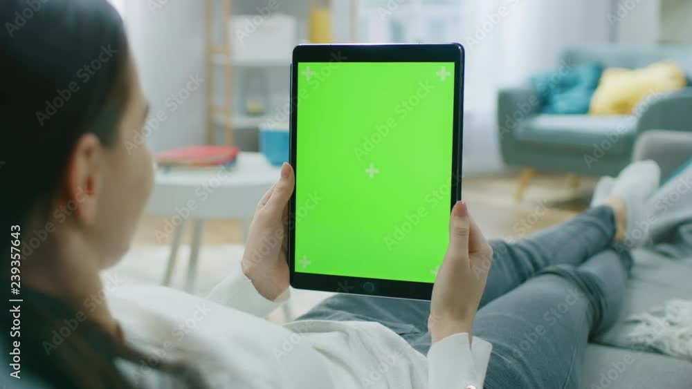 Poster young woman at home resting on a couch using with green mock-up screen tablet computer in vertical p