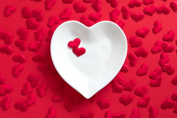 Elegant composition for Valentine's day. White plate in shape of heart with red satin heart on red background. Valentine day concept for design. Flat lay, top view, copy space