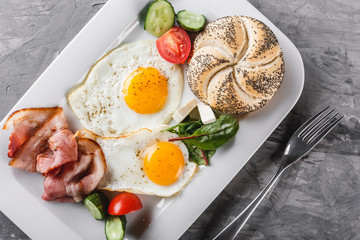 Breakfast, fried eggs, bun, bacon, prosciutto, fresh salad on plate on grey table surface. Healthy food