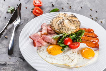 Breakfast, fried eggs, bun, sausage, bacon, prosciutto, fresh salad on plate on grey table surface. Healthy food