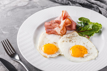 Breakfast, fried eggs, bacon, prosciutto, fresh salad on plate on grey table surface. Healthy food, top view, flat lay