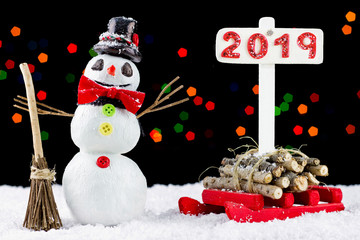 Snowman with a 2019 signpost and red sled on bokeh lights background