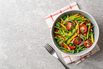 Salad with asparagus, bell pepper, cherry tomatoes, red onion and green peas in ceramic bowl on stone background. Top view. Copy space.