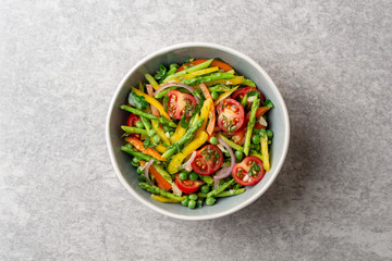 Salad with asparagus, bell pepper, cherry tomatoes, red onion and green peas in ceramic bowl on stone background. Top view. Copy space.
