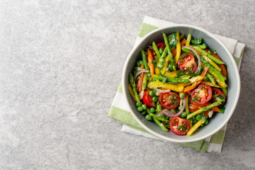 Salad with asparagus, bell pepper, cherry tomatoes, red onion and green peas in ceramic bowl on stone background. Top view. Copy space.