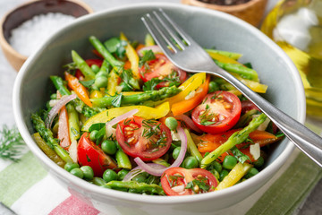 Salad with asparagus, bell pepper, cherry tomatoes, red onion and green peas in ceramic bowl on stone background. Top view. Copy space.