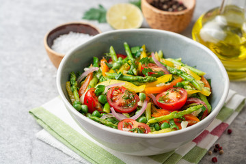 Salad with asparagus, bell pepper, cherry tomatoes, red onion and green peas in ceramic bowl on stone background. Top view. Copy space.