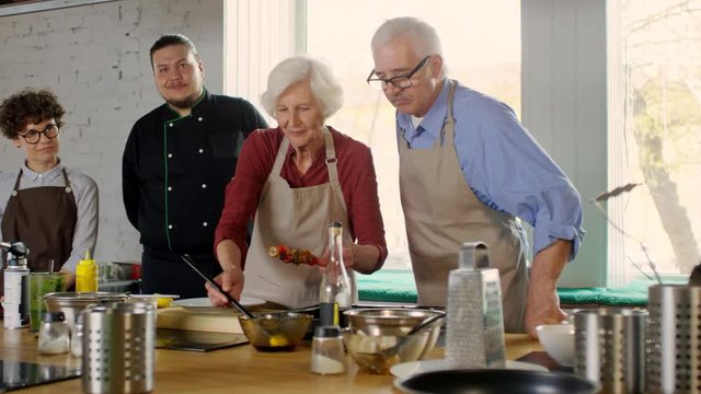Medium shot of old woman cooking vegetarian skewers while other people including chef watching her actions