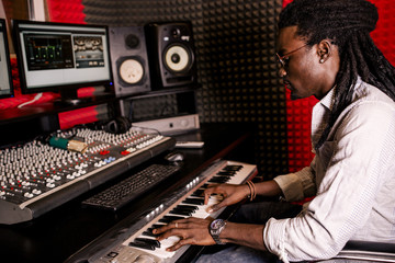 African male sound engineer at work sitting at the mixing console playing on the synthesizer side view