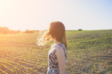Girl in rustic dress is standing in the field with seed beds. Beautiful woman is turning her head in the sunset lights. Wind in her hair. Eco friendly agriculture modern ideas.