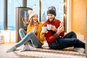 Young couple dressed in bright sweaters sitting together with hot drinks near the fireplace in the...