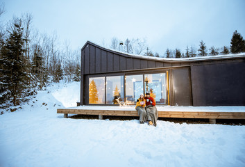 Young couple warming up with plaid and hot drinks sitting on the terrace of the modern house in the mountains durnig the winter holidays. Wide landsacpe view