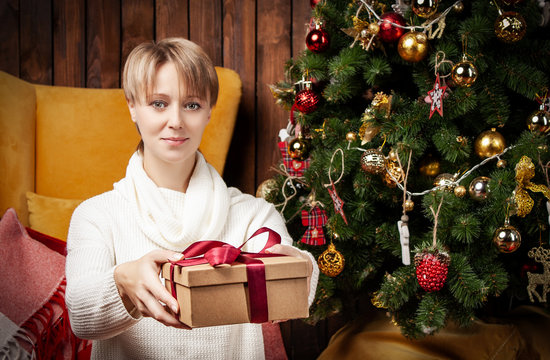 Beautiful woman wearing a warm sweater and socks, sitting near tha Christmas tree holding gift boxes