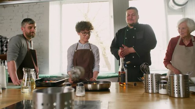 Pan shot of woman holding cooking tongs in her hand, frying some food in pan while on background chef is talking and others listening