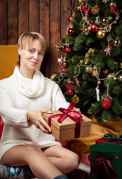 Beautiful woman wearing a warm sweater and socks, sitting near tha Christmas tree holding gift boxes