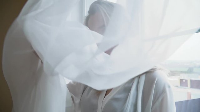 Portrait of beautiful woman with pierced nose close up. Female holds tulle and then lowers it, covering herself. A girl in a nightie standing near a large bright window in the bedroom.