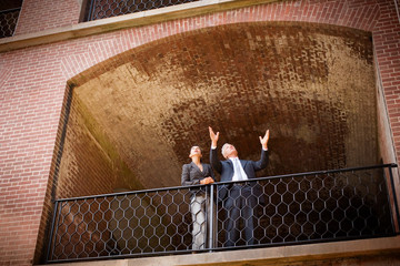 Mature businessman gesturing while standing next to a female colleague on the balcony of a brick building.