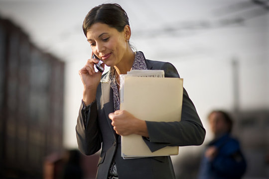 Mid-adult businesswoman listening on her cell phone while holding a folder of documents in the city.