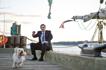 Suited businessman smiling while watching his small white dog run along a wooden wharf.