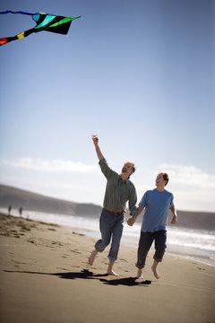 Homosexual male couple flying a kite while holding hands on a beach.