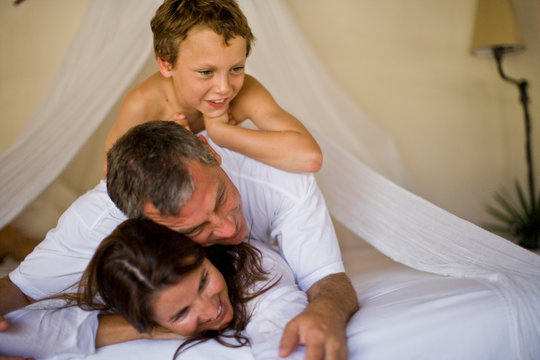 Boy lying on top of a mid-adult couple on a bed.