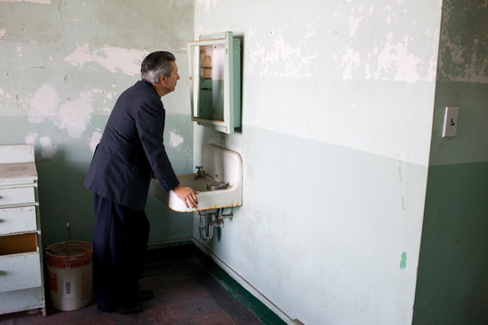 Mature adult businessman leaning on a sink while looking in an old cabinet in a derelict building.