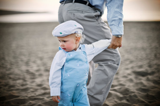 Young toddler wearing a flat cap while holding hands and walking along a sandy beach.
