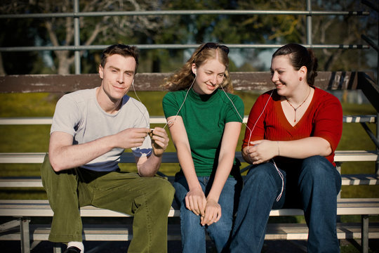 Three teenagers sharing headphones to listen to music on an MP3 player while sitting in bleachers outdoors in the sun.
