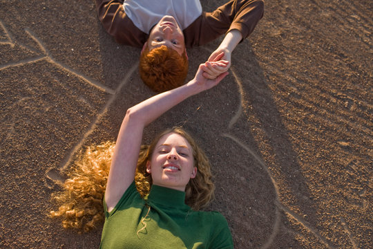 Portrait of a smiling teenage couple holding hands lying on their backs on a heart shape drawn in the dirt.