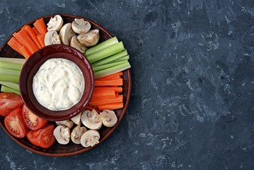 Vegetable tray with a ranch dip on a dark background. Raw vegetable mix includes sliced mushrooms, carrots, celery, tomatoes.