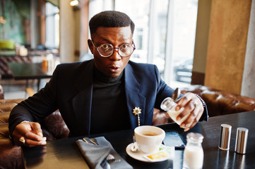 Fashionable african american man in suit and glasses pours cream into coffee at cafe.