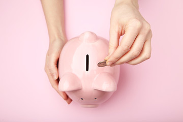 Female hand putting coin into piggy bank on pink background