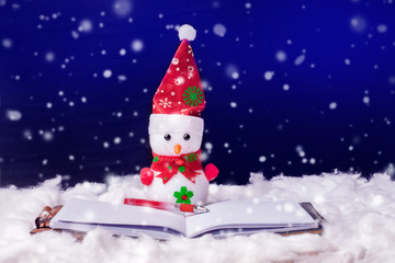 Toy snowman in front of an open book. Fiction reading. Snowman on a dark background during the snowfall. Happy Christmas and New Year's greetings_