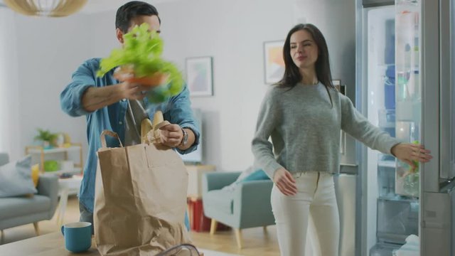 Beautiful Young Couple Come to the Kitchen with Fresh Groceries in Brown Paper Bag. Man is Handing Fresh Salad Greens, Apples and Oranges to the Girl Who Puts Them in the Fridge. 