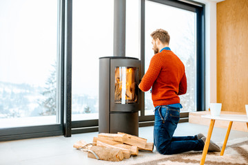 Handsome man dressed in sweater kindling fireplace in the modern house during the winter