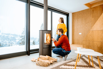 Young couple dressed in bright sweaters warming up near the fireplace in the modern house in the mountains during the winter - obrazy, fototapety, plakaty