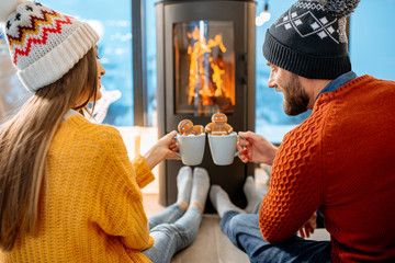 Young couple dressed in bright sweaters with hot drinks near the fireplace in the modern house in the mountains durnig winter time