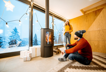 Young couple dressed in bright sweaters sitting near the fireplace in the modern house in the...