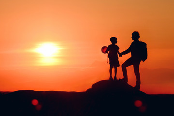 Dad and boy with balloon on fathers day travel together in mountains, beautiful sunset background. Father and son hold hands and look at each other, idyllic scene in evening landscape with setting sun