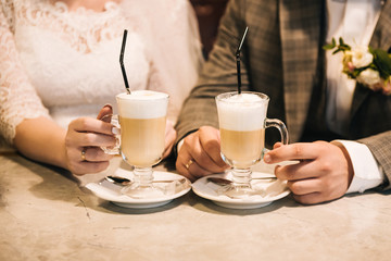 Latte cups in the hands of the newlyweds. The bride and groom drink coffee in a cafe