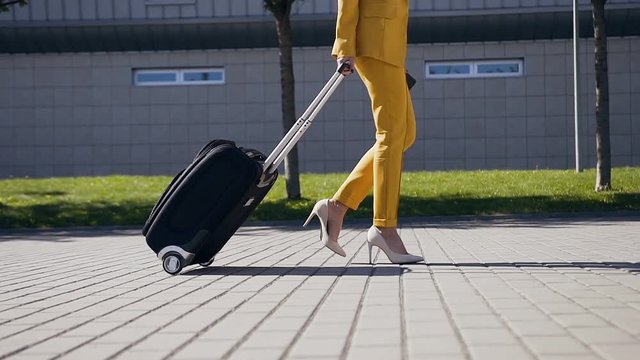 Slim business woman in stylish suit pulls a Suitcase, hurries to a business meeting. Attractive business woman going on a business trip pulling her suitcase along the sidewalk behind her of her legs
