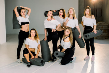 Fototapeta premium Group portrait of young sporty excited beautiful girls with exercise yogs mats standing in modern gym with large window, laughing and talking together.