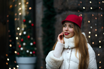 lovely blonde woman with red lips wearing funny hat and winter coat walking down the street decorated with garlands. Space for text