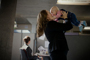 Young businesswoman holding her young baby aloft and touching noses while standing inside an office.