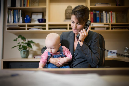 Mid-adult businesswoman talking on the phone and holding her young baby while seated at a desk in her office.