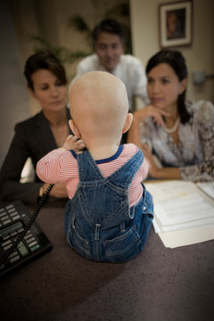 Young baby sitting on a desk in front of three businesspeople inside an office.
