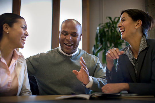 Three business colleagues laughing while sitting at a table during a meeting inside a boardroom.